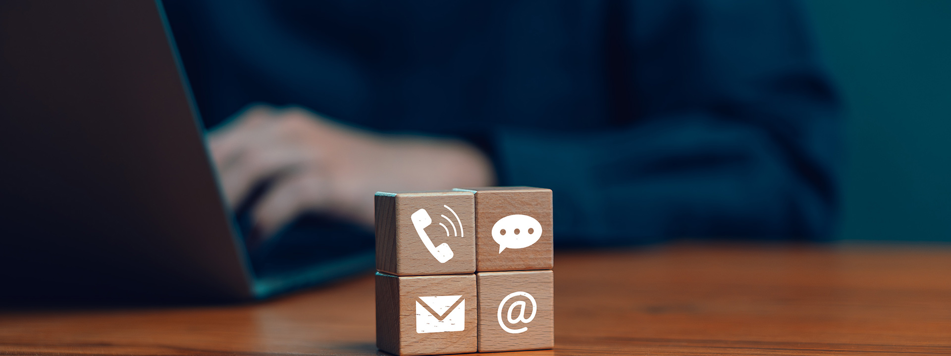 Blurred person's hand in the background typing on a laptop, 4 wooden blocks stacked on each other with icons representing a telephone, email, website and chat.