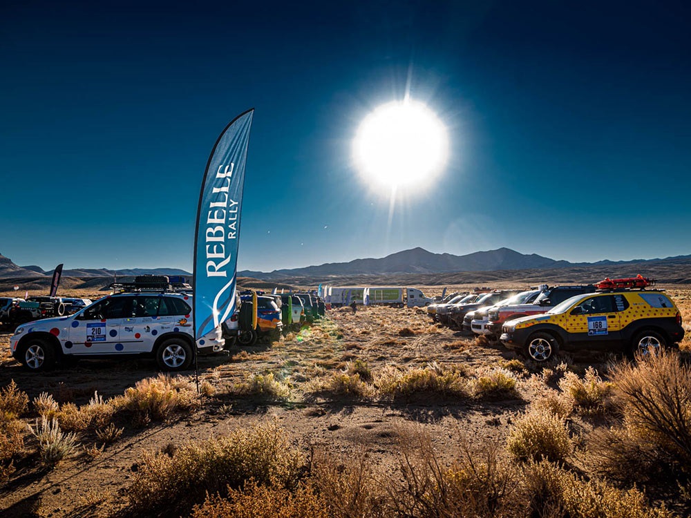 Group of vehicle parked in a field for Rebelle Rally with the sun shining brightly in the distance