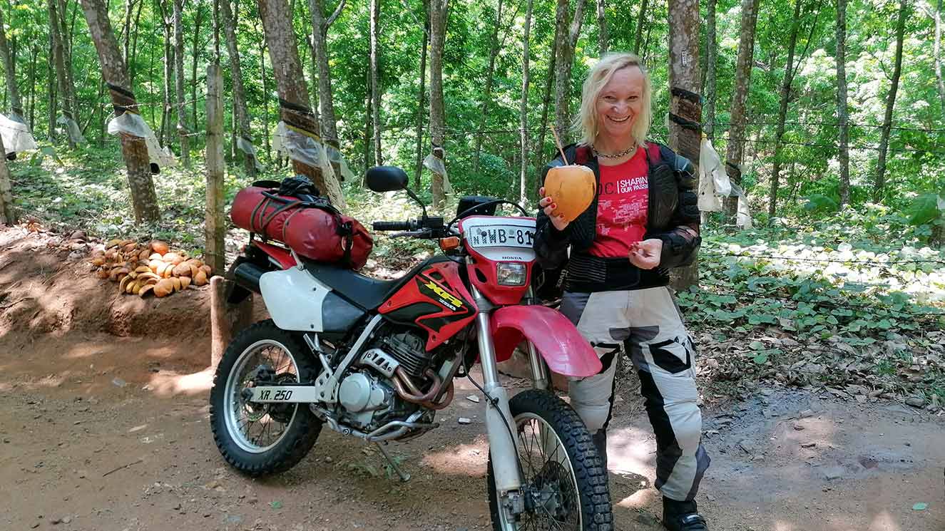 Rebecca Kleis drinking from a coconut with motorcycle in wooded area