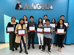 Group of standing together in front of a Magna logo holding certificates