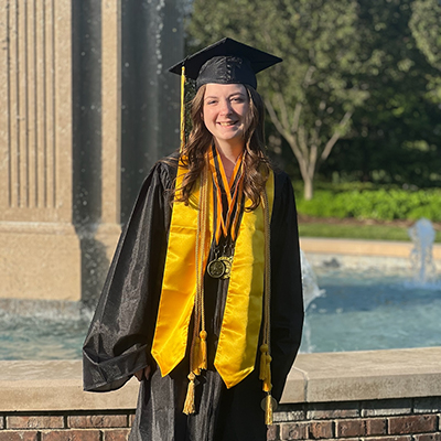 Erin Craft standing in front of a fountain wearing graduation cap and gown