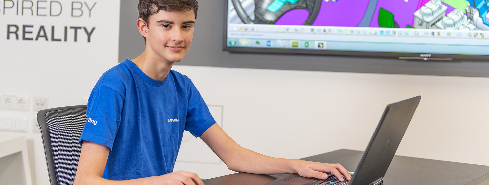 Young worker in a blue shirt using a laptop with a 3D design projected on a screen behind