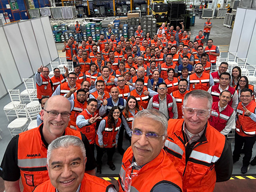 Miguel Arellano & top management of Magna with factory workers in bright orange and red safety vests. Smiling, they gather in a manufacturing facility for a team photo, radiating camaraderie and teamwork.