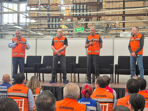 Miguel Arellano and Magna top management in orange safety vests stand on a stage addressing an audience in a manufacturing facility. The audience also wears similar vests.