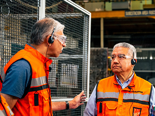 Miguel Arellano and Swamy Kotagiri in safety gear and headsets have a serious discussion in a manufacturing facility. Both wear orange vests and safety glasses, indicating a focus on safety.
