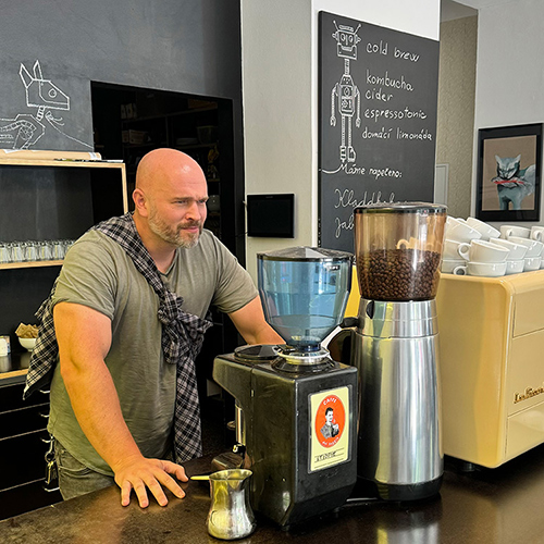 Miroslav Frontz with a plaid shirt over his shoulder stands at a coffee bar, leaning on the counter. A chalkboard menu and coffee machine are visible. The mood is relaxed.