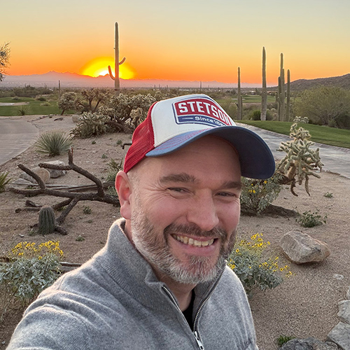 Miroslav Frontz in a red and white cap stands in a desert landscape at sunset. Cacti and shrubs are visible, with a warm, colorful sky in the background.