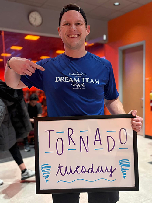 青い「Make-A-Wish Dream Team」シャツを着て「Tornado Tuesday」と書かれた看板を持つ男性